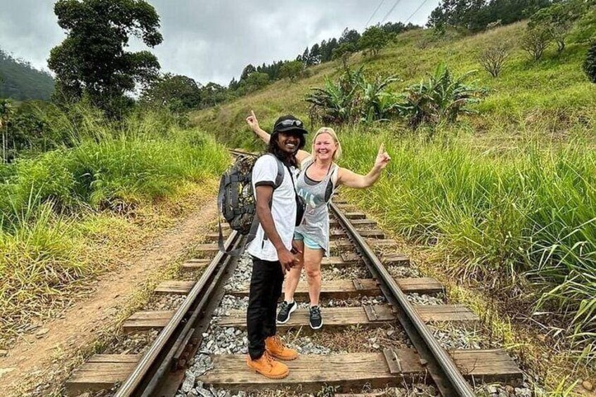 Two people standing on railway tracks with greenery and hills in the background. 3