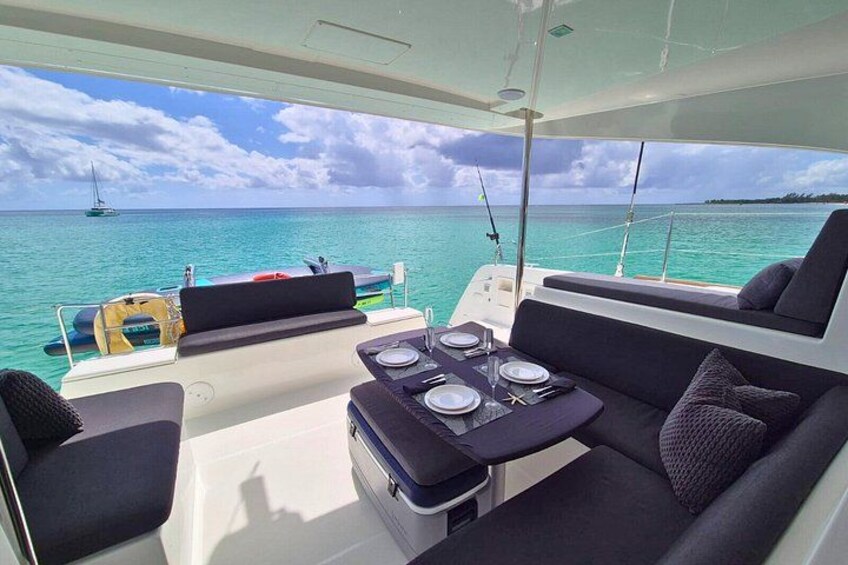 A boat deck with a table set for dining, a cushioned seating area, and a view of the ocean with another boat in the distance. 3