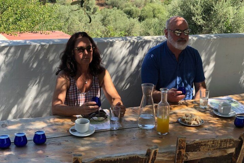 Two people sitting at an outdoor table with drinks and food, surrounded by greenery. 2