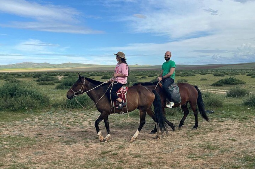 Two people riding horses on a dirt path in a grassy field with distant hills. 4