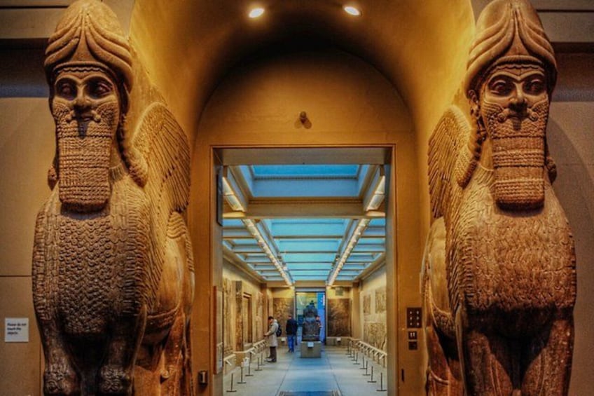 A hallway with two large stone statues on either side, leading to a bright interior space. 2
