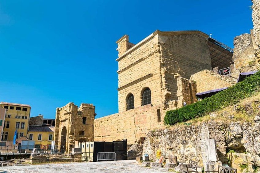 A historic stone building with arched windows and a tower, surrounded by modern buildings and a clear blue sky. 4