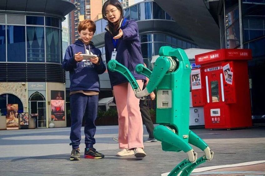 Two people interacting with a green robotic device in an urban setting with modern architecture and a red mailbox in the background. 1