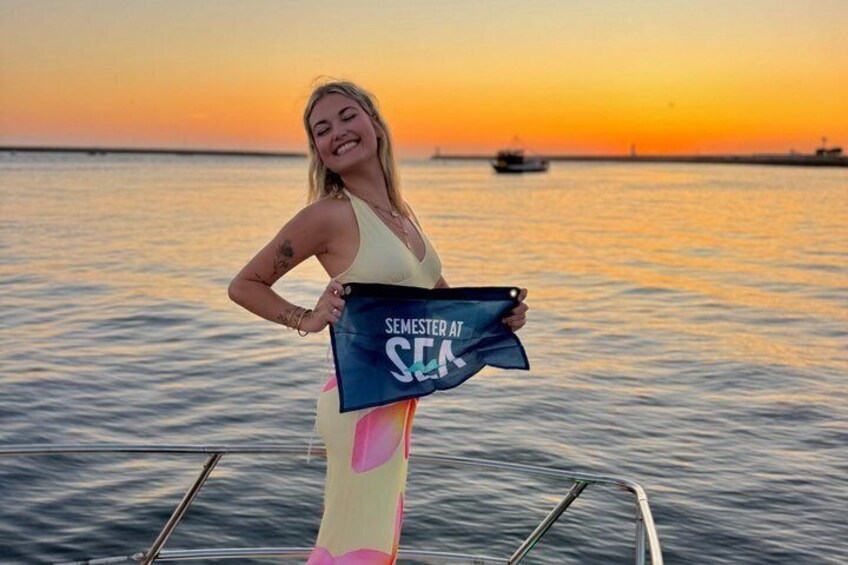 A person holding a bag with the text "SEMESTER AT SEA" on a boat at sunset. 1