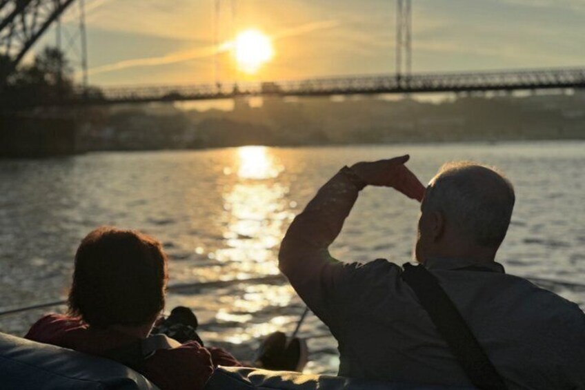 Two people on a boat, one pointing towards the sunset over a body of water with a bridge in the background. 3