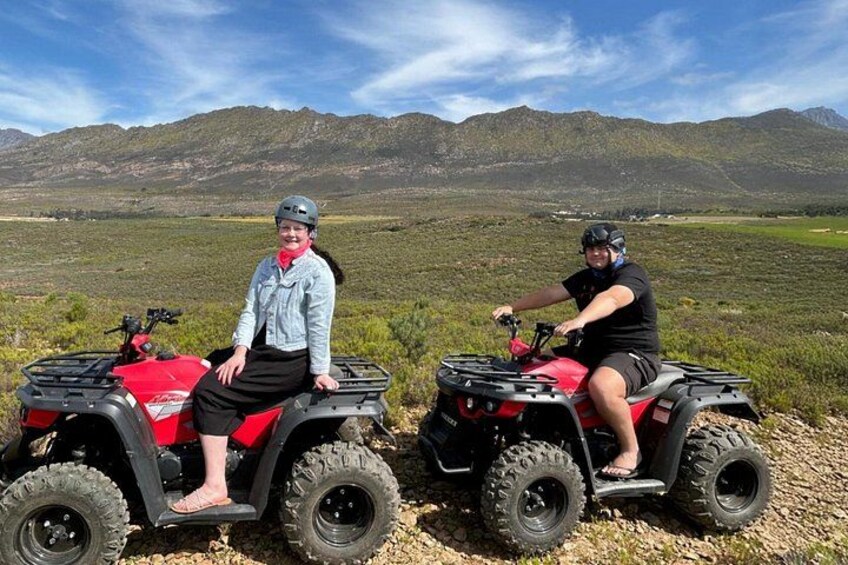 Two people on red ATVs in a grassy field with mountains in the background. 2