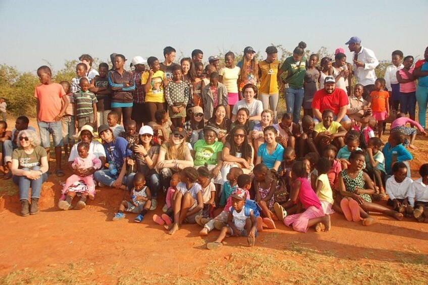 A group of people gathered outdoors, some sitting and some standing, in a dry, reddish terrain with sparse vegetation. 2