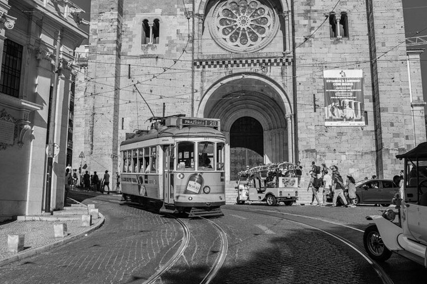 A vintage tram on a cobblestone street, a historic building with a large arched doorway, and pedestrians walking by. 4