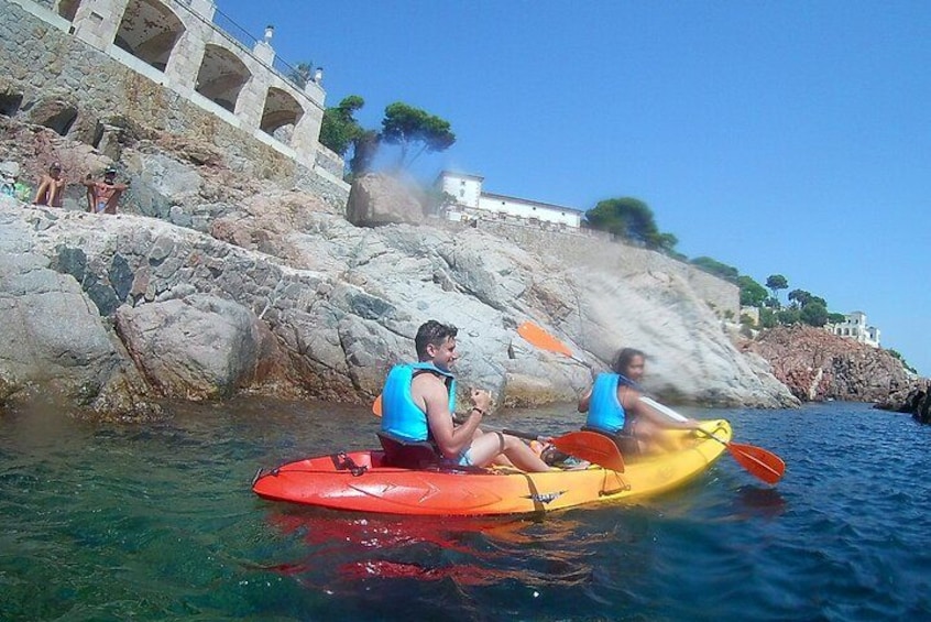 Two people kayaking on a calm body of water with rocky cliffs and a white building in the background. 4