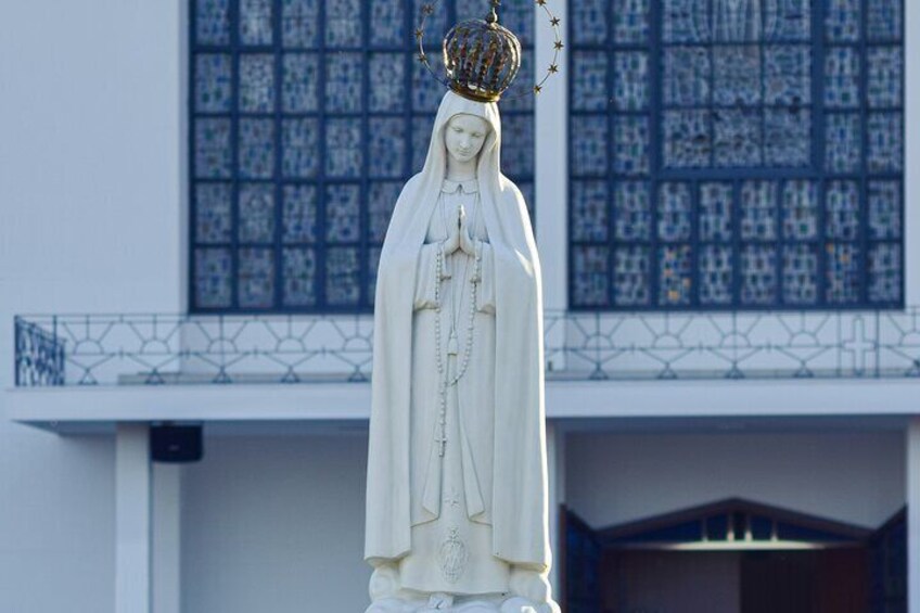 Une statue représentant une figure drapée, coiffée d’une couronne, se dressant devant un bâtiment orné d’une fenêtre à motifs. 1