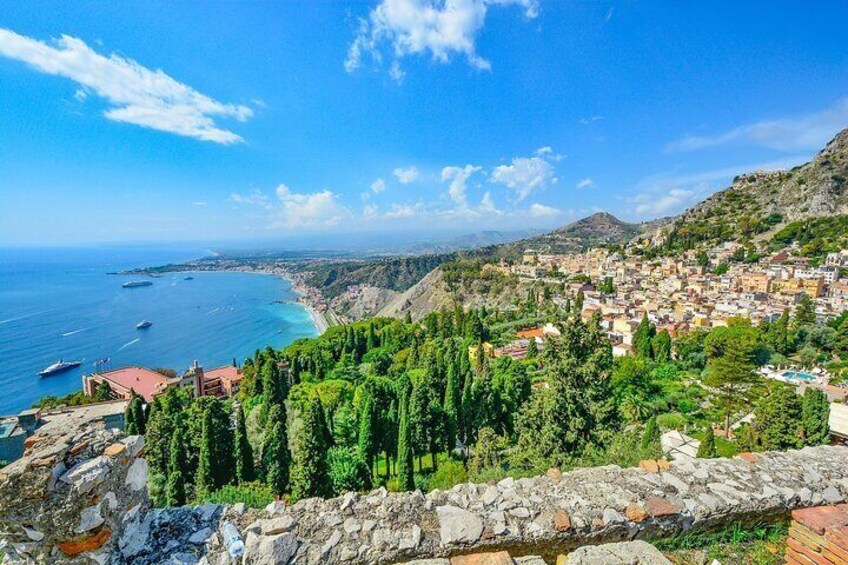 A coastal town with a stone wall, green trees, and boats on the sea. 1