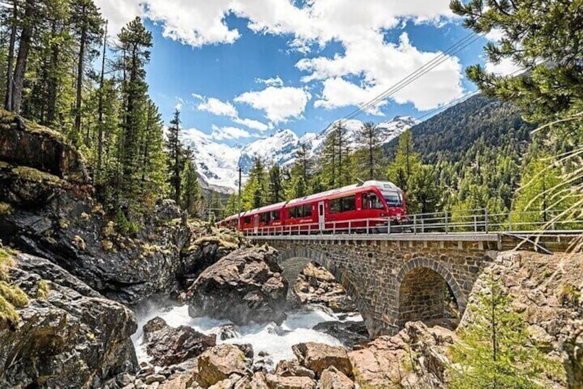 Un treno rosso attraversa un ponte di pietra su un torrente roccioso in una zona montuosa e boscosa. 1