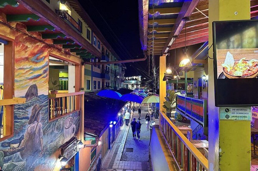 A brightly lit pedestrian walkway with murals, overhead lighting, and a signboard. 2
