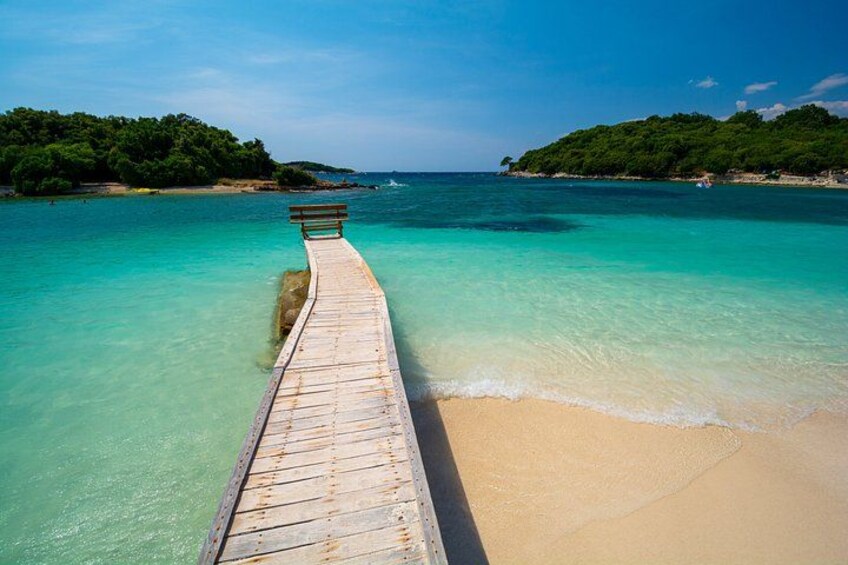Una banchina in legno si protende su acque turchesi, conducendo a una spiaggia sabbiosa con vegetazione rigogliosa sullo sfondo. 1