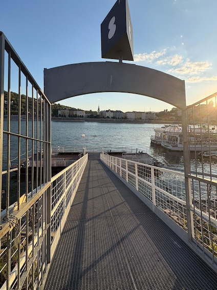 A bridge with a metal railing, a sign with a white symbol, and a view of a river with boats and buildings in the background. 5