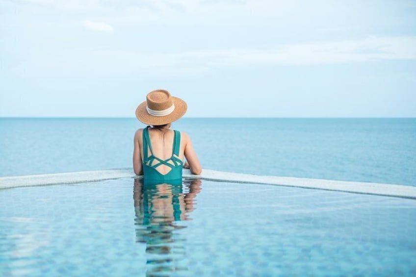 A person in a blue swimsuit and straw hat sitting in a pool with a view of the ocean. 1