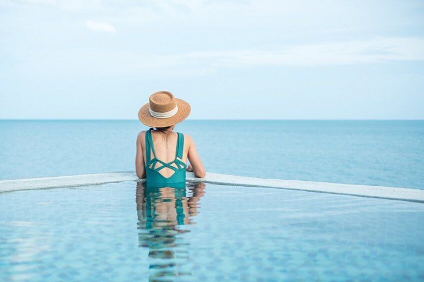A person in a blue swimsuit and straw hat sitting in a pool with a view of the ocean. 1
