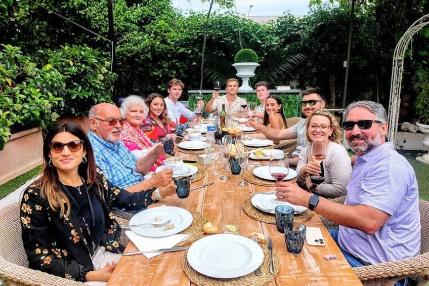 A group of people seated around a wooden table outdoors, engaged in a meal with plates, glasses, and cutlery. 2