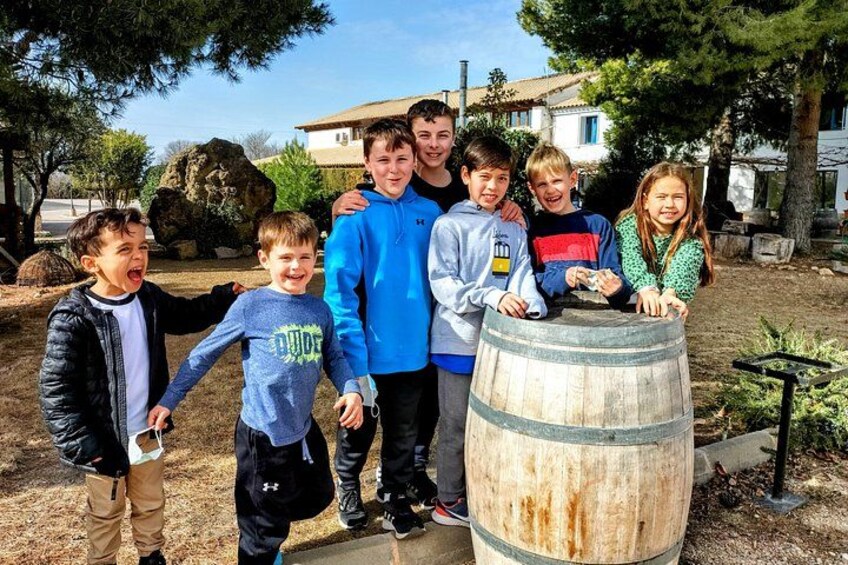 A group of people standing around a wooden barrel in an outdoor setting with trees and a building in the background. 5