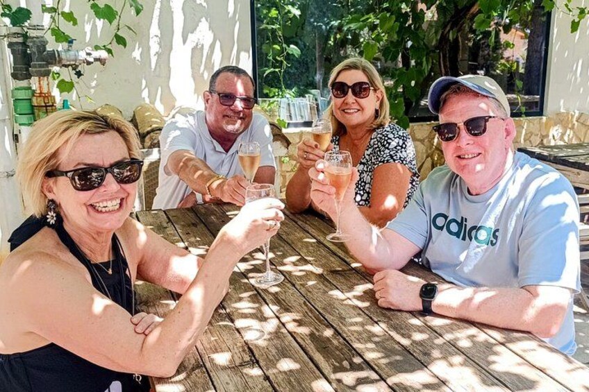 Four people sitting at a wooden table outdoors, holding glasses of a beverage. 1