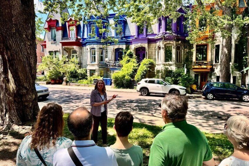 A person gesturing to a group of people on a street with colorful Victorian-style houses. 1