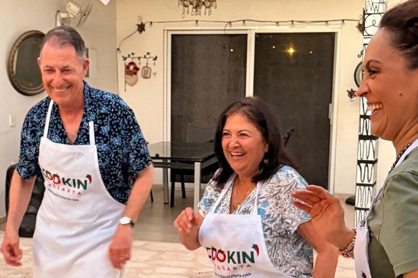 Tres personas con mandiles que llevan el logo 'Cookin' ALERTA', de pie en una habitación con una mesa y sillas para comer. 4