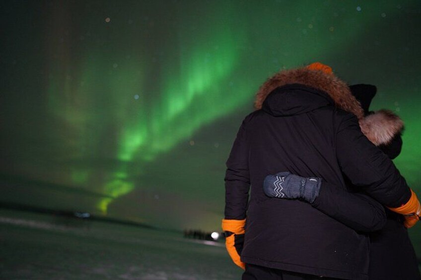 A person wearing a black jacket with a fur-lined hood and orange accents, standing outdoors at night. 1