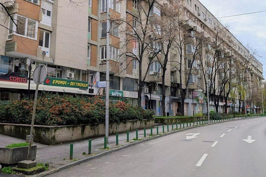 A street lined with multi-story buildings, a sidewalk with green railings, and a road with directional arrows. 5