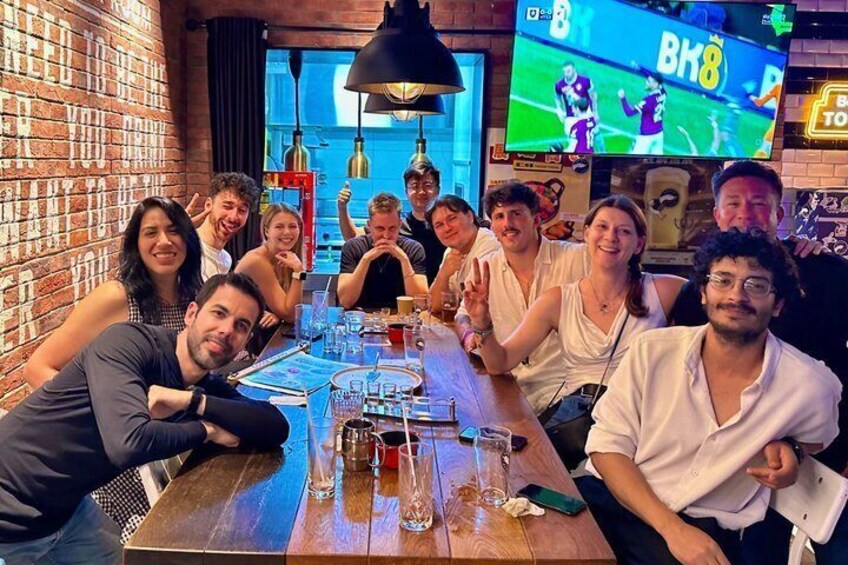 A group of people seated around a wooden table in a bar with a brick wall and a TV screen displaying a sports event. 5