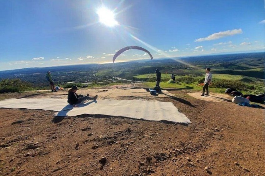 En grupp människor på en lerig åker, med en fallskärm på himlen, en klarblå himmel och ett avlägset landskap i bakgrunden. 2