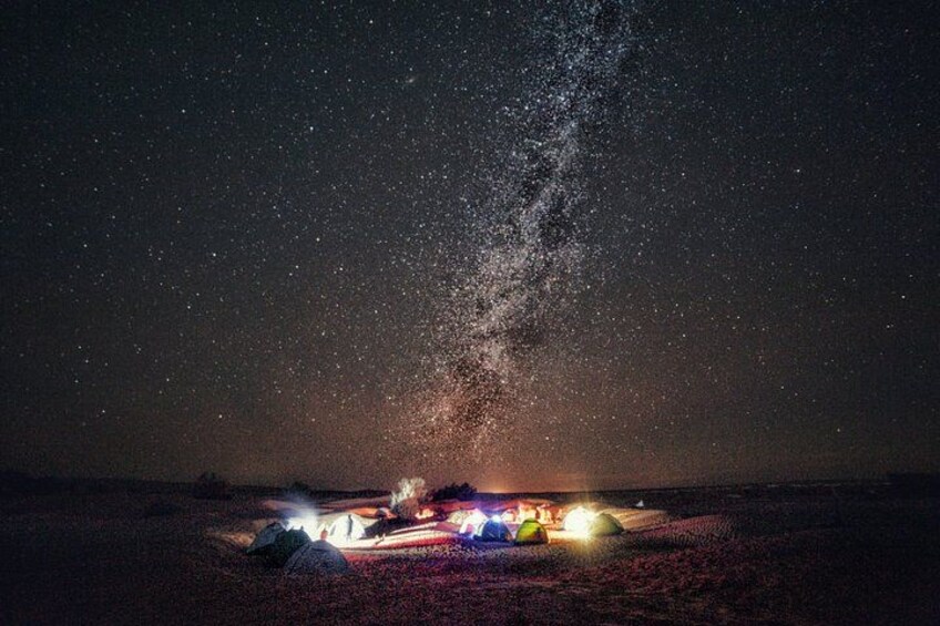 Cielo nocturno estrellado con la Vía Láctea visible, un camping con tiendas de campaña y una fuente de luz distante. 3