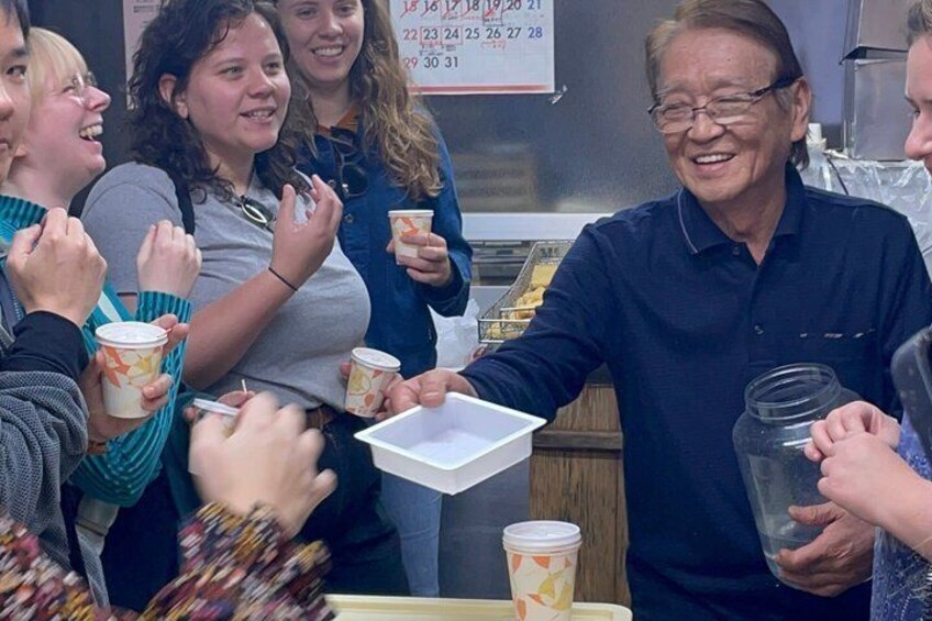 Un grupo de personas en un puesto de comida, algunas sosteniendo vasos y una persona repartiendo una bandeja. 2