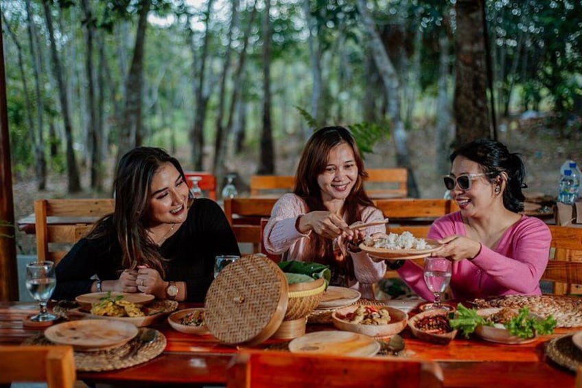 Three people dining outdoors with a variety of dishes and a forested background. 2