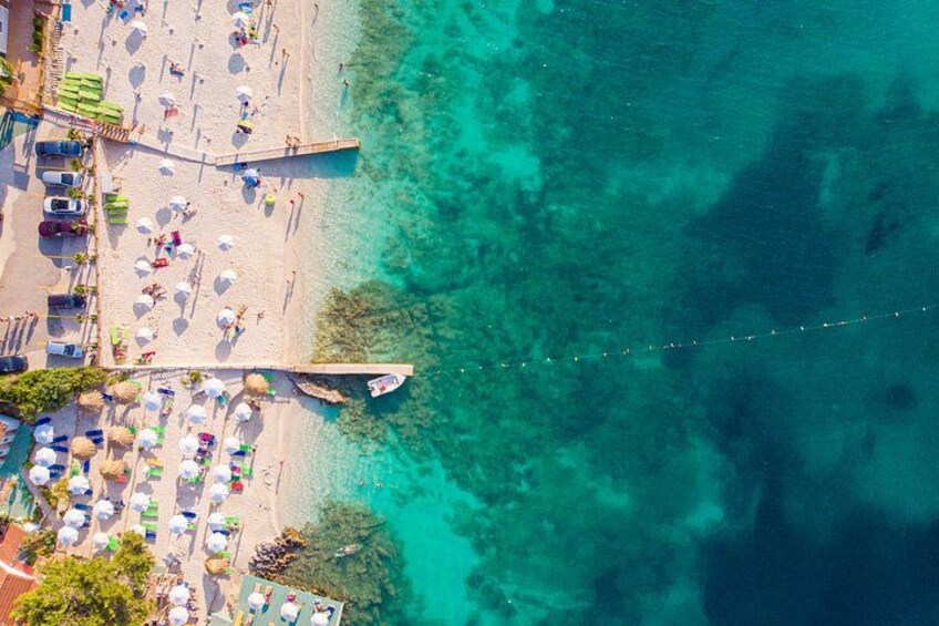 Luftaufnahme eines Strandes mit klarem türkisblauem Wasser, Sonnenschirmen und Personen. 2