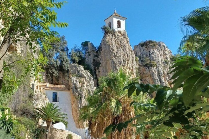 A white church with a bell tower on a rocky cliff, surrounded by greenery. 1
