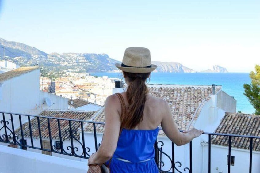 A person in a blue dress and hat overlooking a coastal town with buildings and a view of the sea. 2