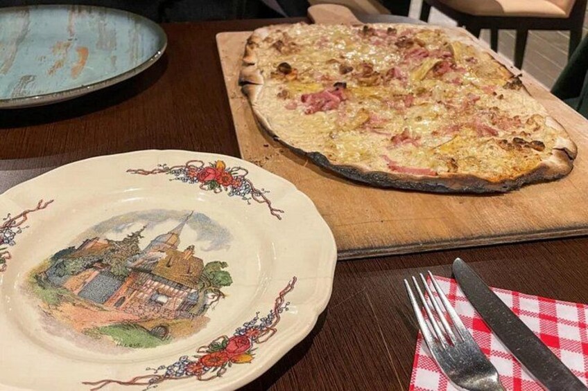 A wooden table with a pizza on a wooden board and a decorative plate with a building illustration. 1