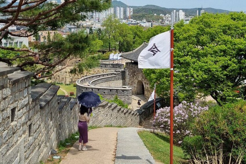 A stone pathway leading to a traditional Korean gate, surrounded by greenery and a cityscape in the background. 4