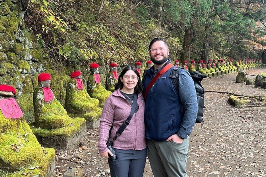 A person standing in front of moss-covered statues with red hats, a forested area with rocks and fallen leaves. 3