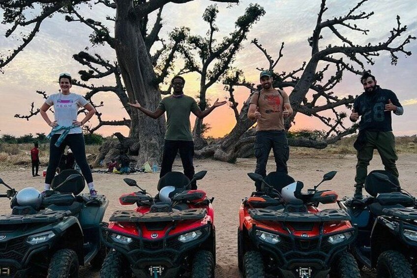 Four people standing with ATVs in a desert landscape with a large tree and a sunset in the background. 3