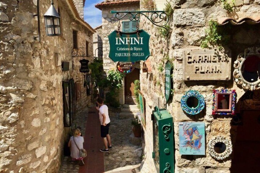 Une rue étroite bordée d’immeubles en pierre, une enseigne indiquant « INFINI, créations de bijoux et parures – perles », et des miroirs décoratifs. 2