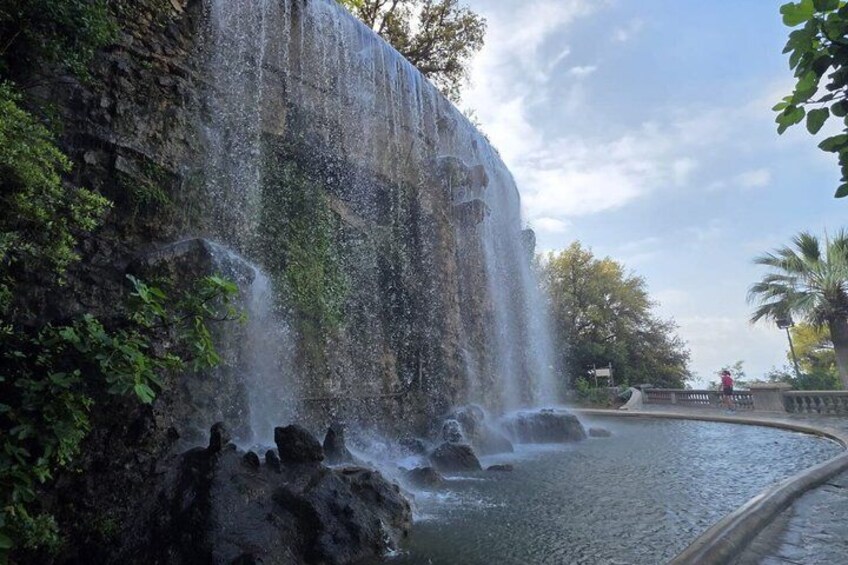 A waterfall cascading over a rocky cliff into a pool, surrounded by greenery and a clear sky. 2