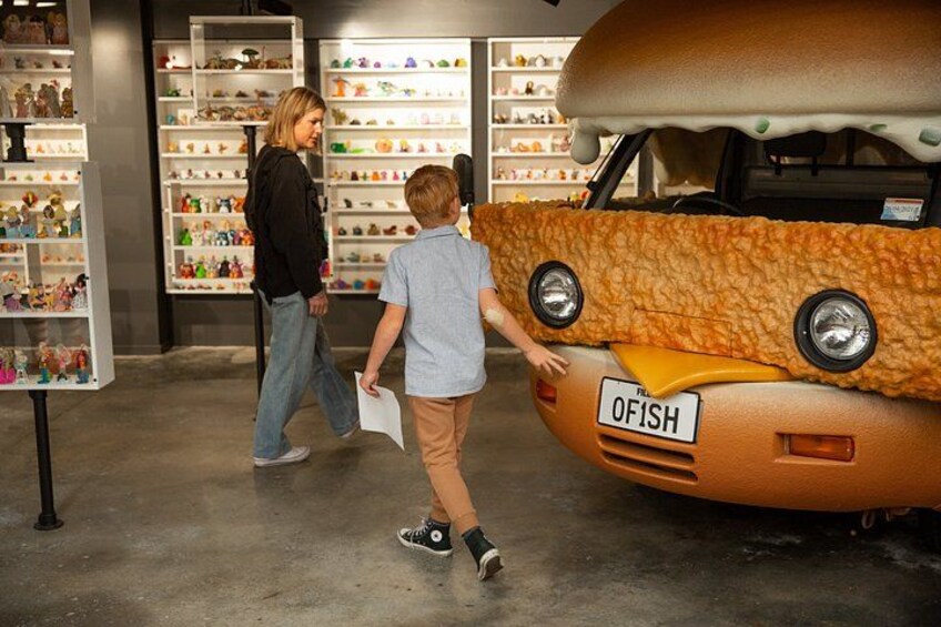 A person walking past a large, orange, fish-shaped vehicle with a license plate reading 'OFISH' in a store with shelves of toys. 2