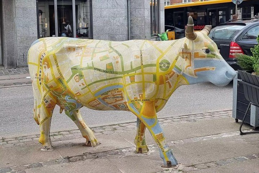 A large cow statue with a map pattern, standing on a street with buildings and cars in the background. 3