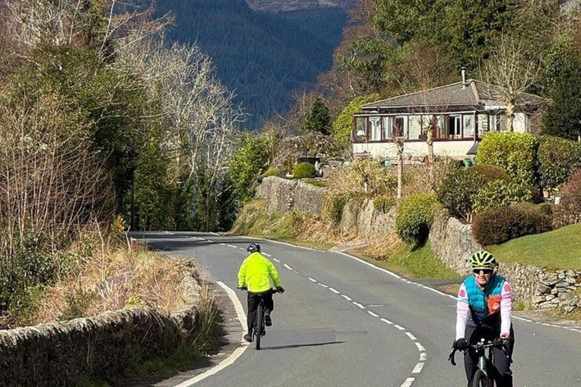 Two cyclists on a winding road with a house and mountains in the background. 5
