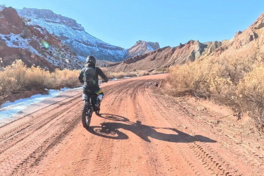 A person riding a motorcycle on a dirt road with snow patches and red rock formations in the background. 2
