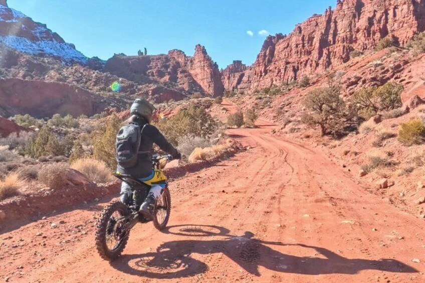 A person riding a dirt bike on a red dirt road in a desert landscape with red rock formations and sparse vegetation. 3