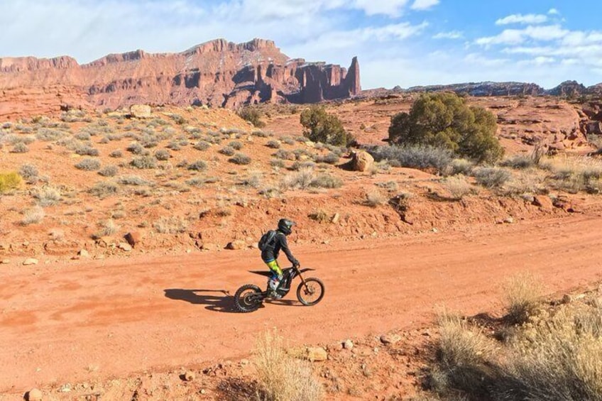 A person riding a mountain bike on a dirt road in a desert landscape with red rock formations in the background. 4