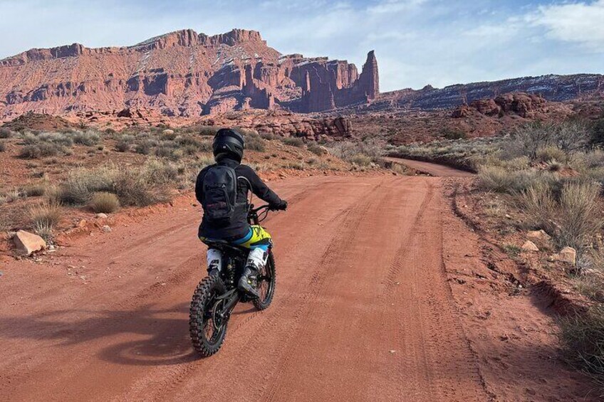 A person riding a dirt bike on a dirt road in a desert landscape with red rock formations in the background. 1