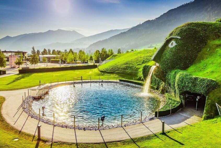 A circular pool with a waterfall, surrounded by green grass and a mountainous landscape. 2